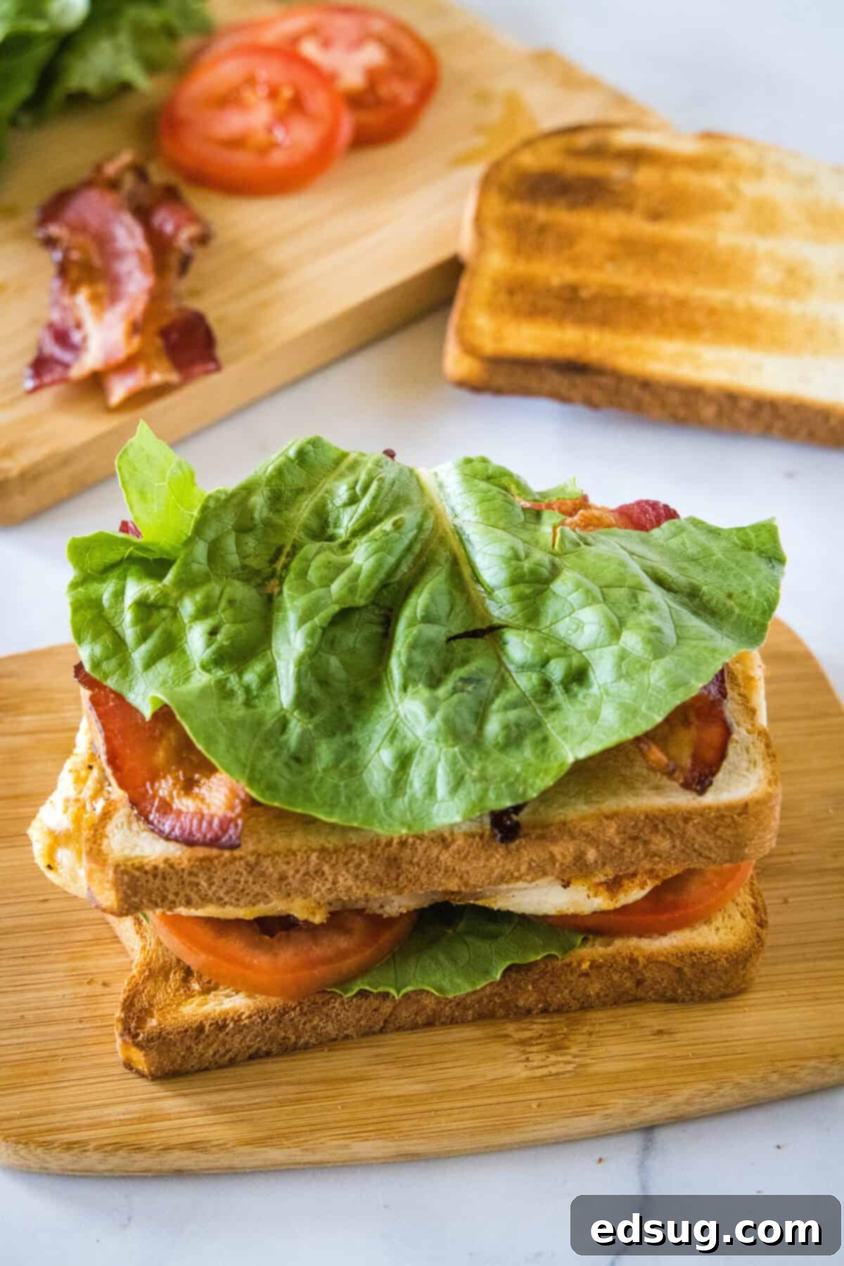 Adding the second toasted bread slice, crispy bacon strips, and an additional layer of fresh lettuce to the chicken club sandwich base on a wooden cutting board.