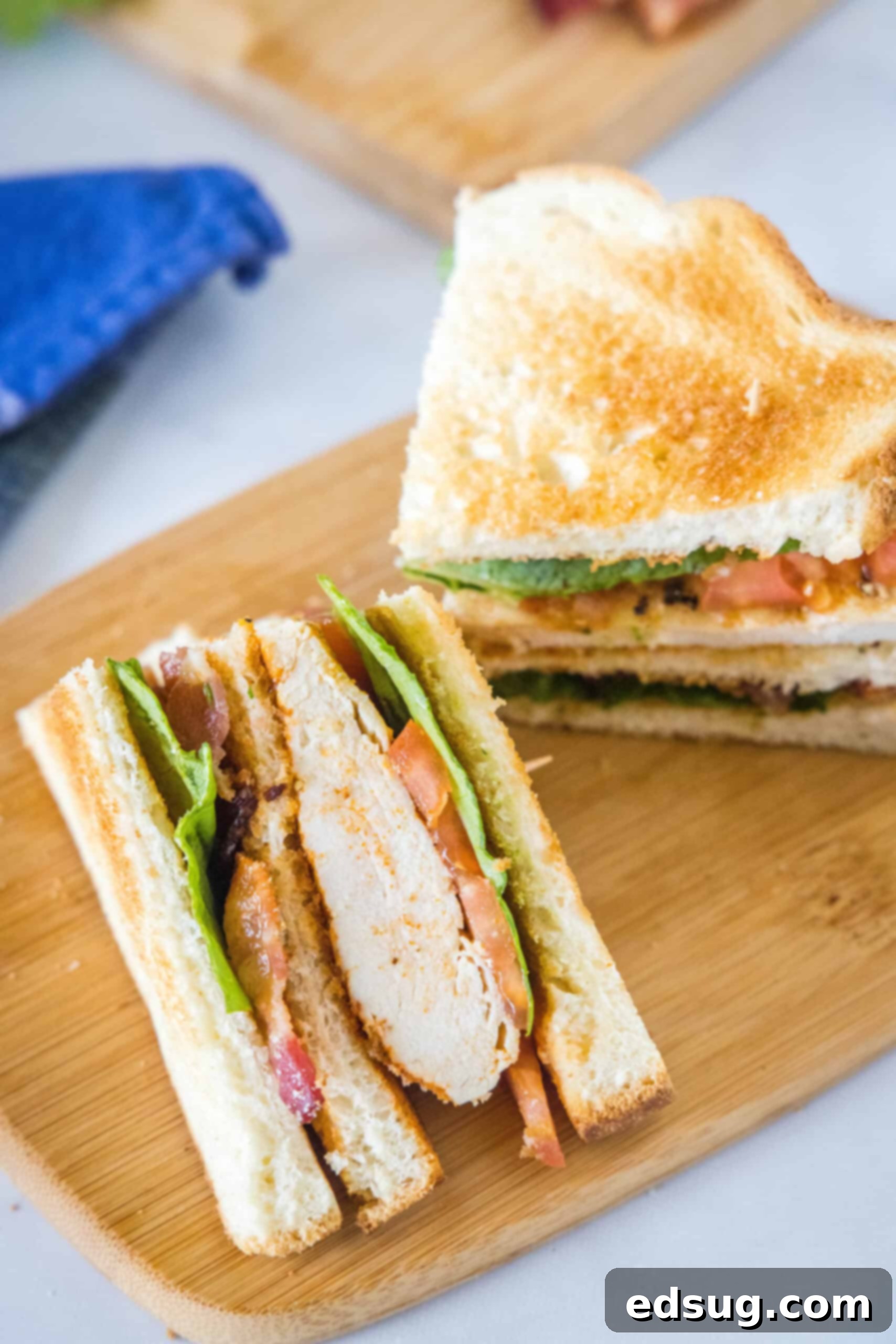 Overhead close-up view of a delicious chicken club sandwich, cut into two perfect halves, showcasing the rich layers of chicken, bacon, lettuce, tomato, and toast, presented on a wooden cutting board.