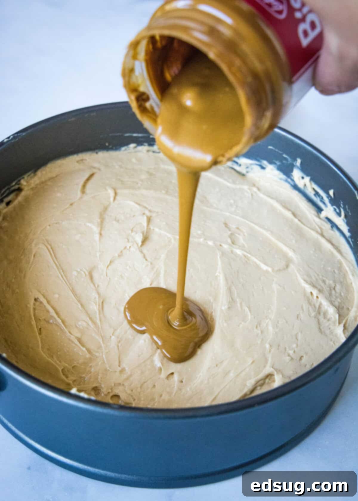 Melted Biscoff spread being drizzled generously over the smooth, light-colored cheesecake filling in a springform pan.