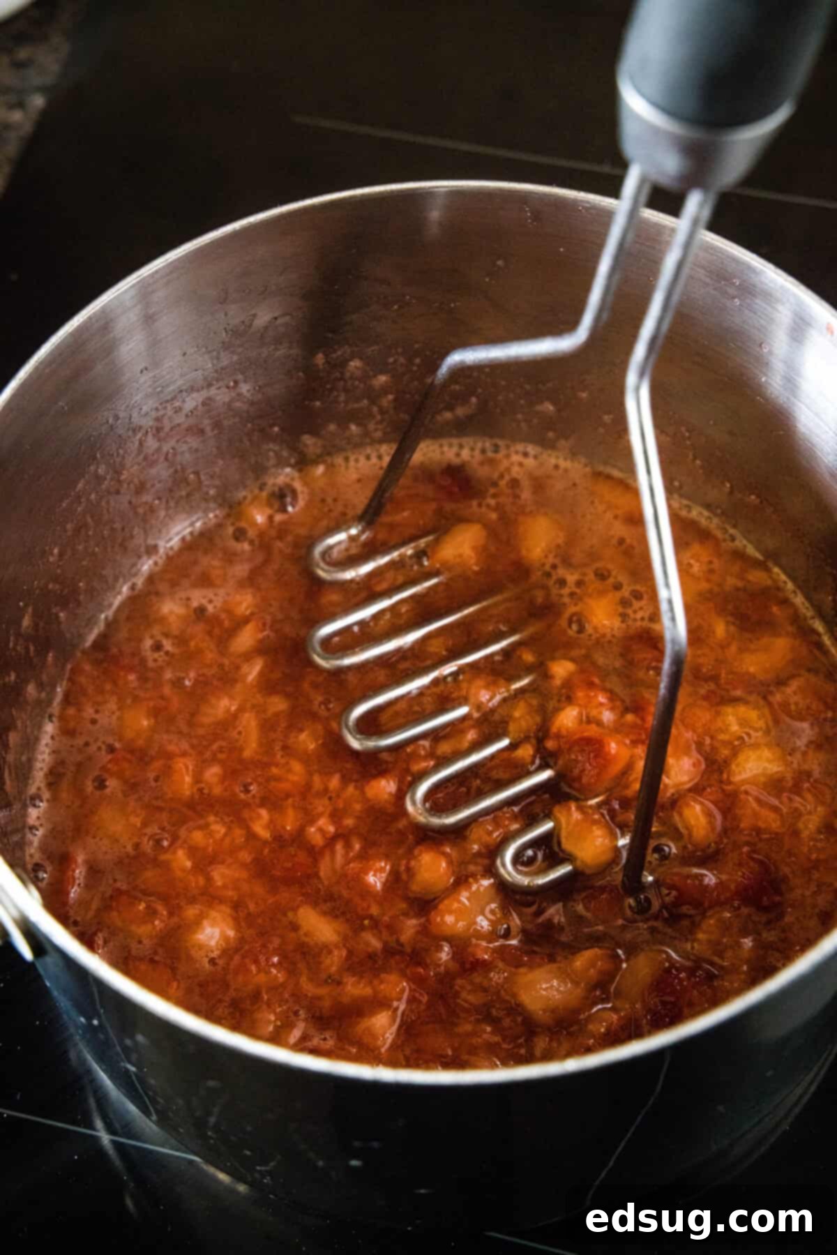 These strawberry cheesecake bars have a tangy cream cheese filling swirled with gooey strawberries, baked on top of a graham cracker crust. A potato masher being used to mash simmered strawberries in a saucepan.