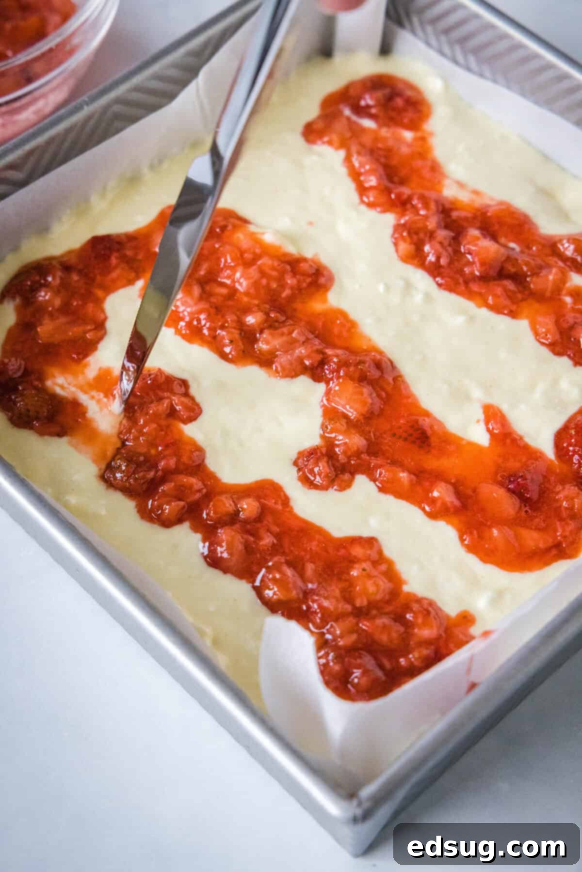 These strawberry cheesecake bars have a tangy cream cheese filling swirled with gooey strawberries, baked on top of a graham cracker crust. A knife being used to swirl strawberry compote into cheesecake filling.