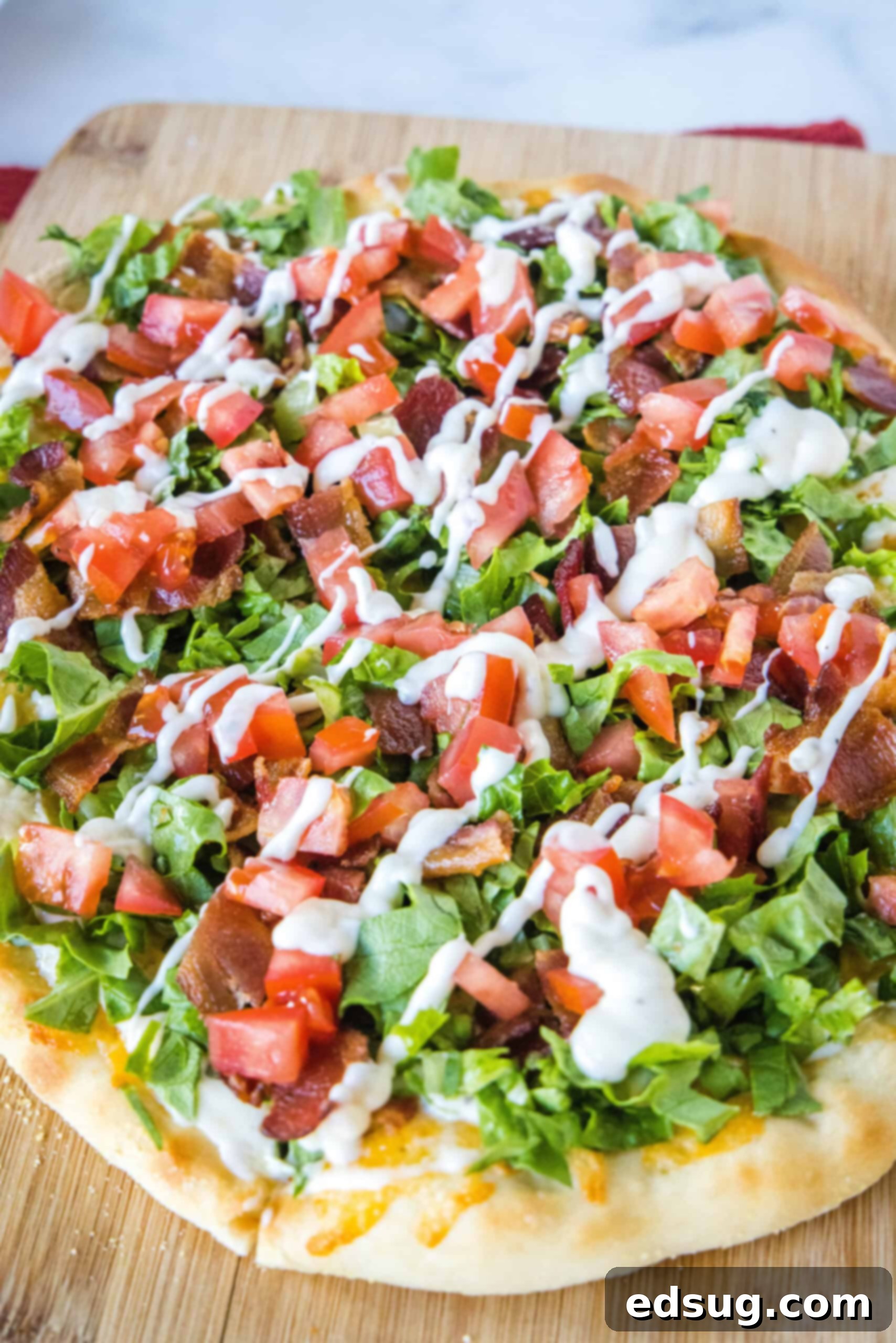 Close up of a BLT pizza topped with a drizzle of aioli on a wooden cutting board.