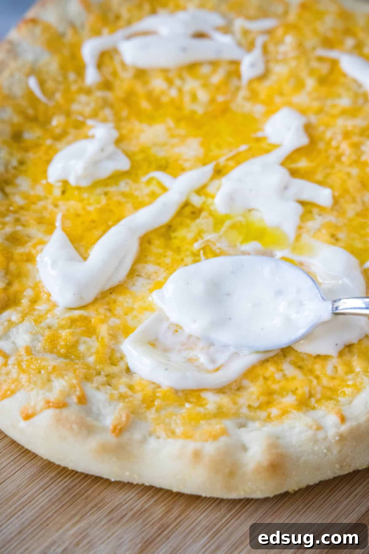 A spoon spreading aioli over melted cheese on top of a baked pizza crust.