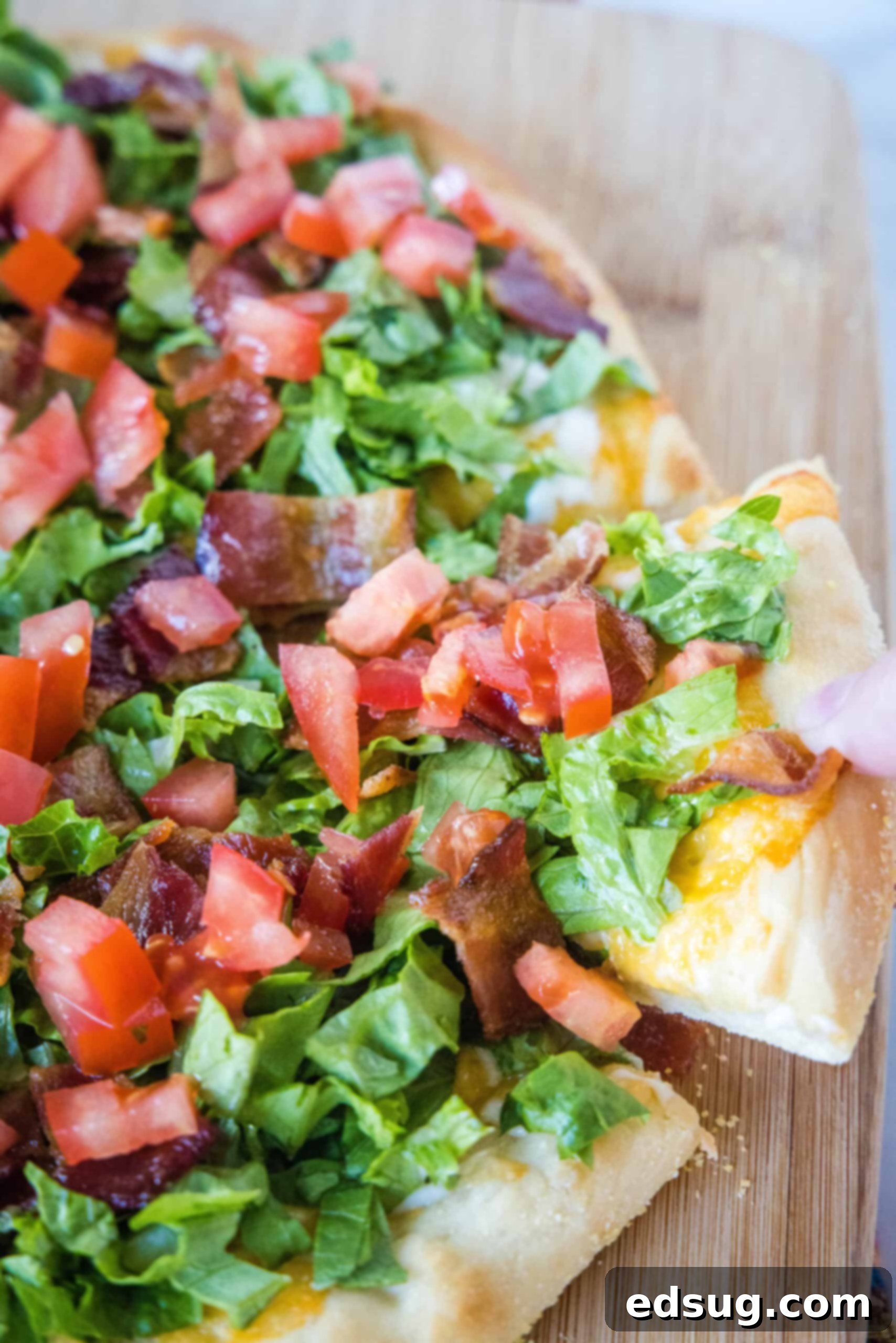 A hand lifting a slice from a BLT pizza on a wooden cutting board.