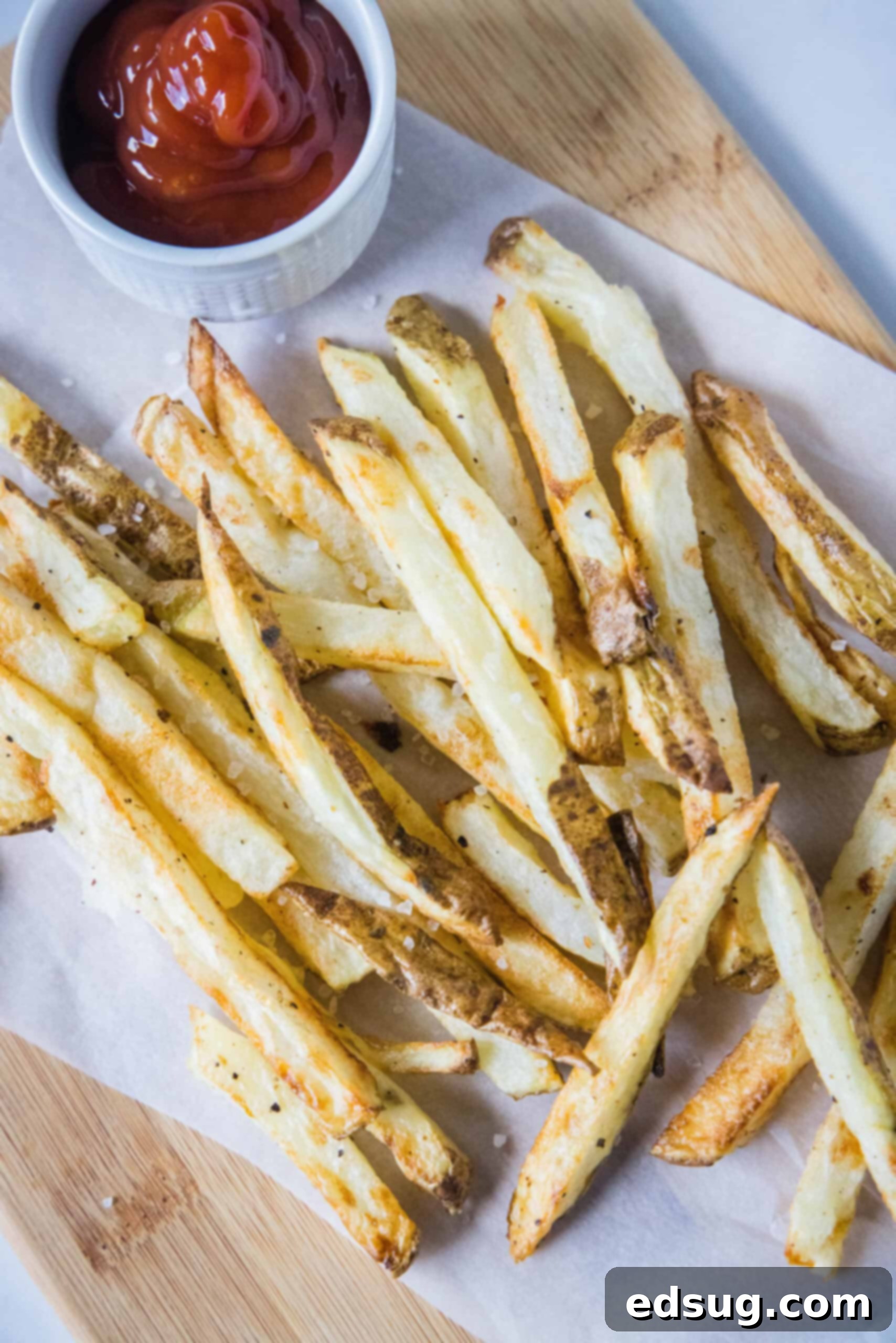 Homemade air fryer French fries are just as crispy as regular fries! Make them in a fraction of the time, with any seasonings you’d like. Overhead view of air fryer French fries on a sheet of parchment paper over a wooden cutting board, next to a small ramekin of ketchup.