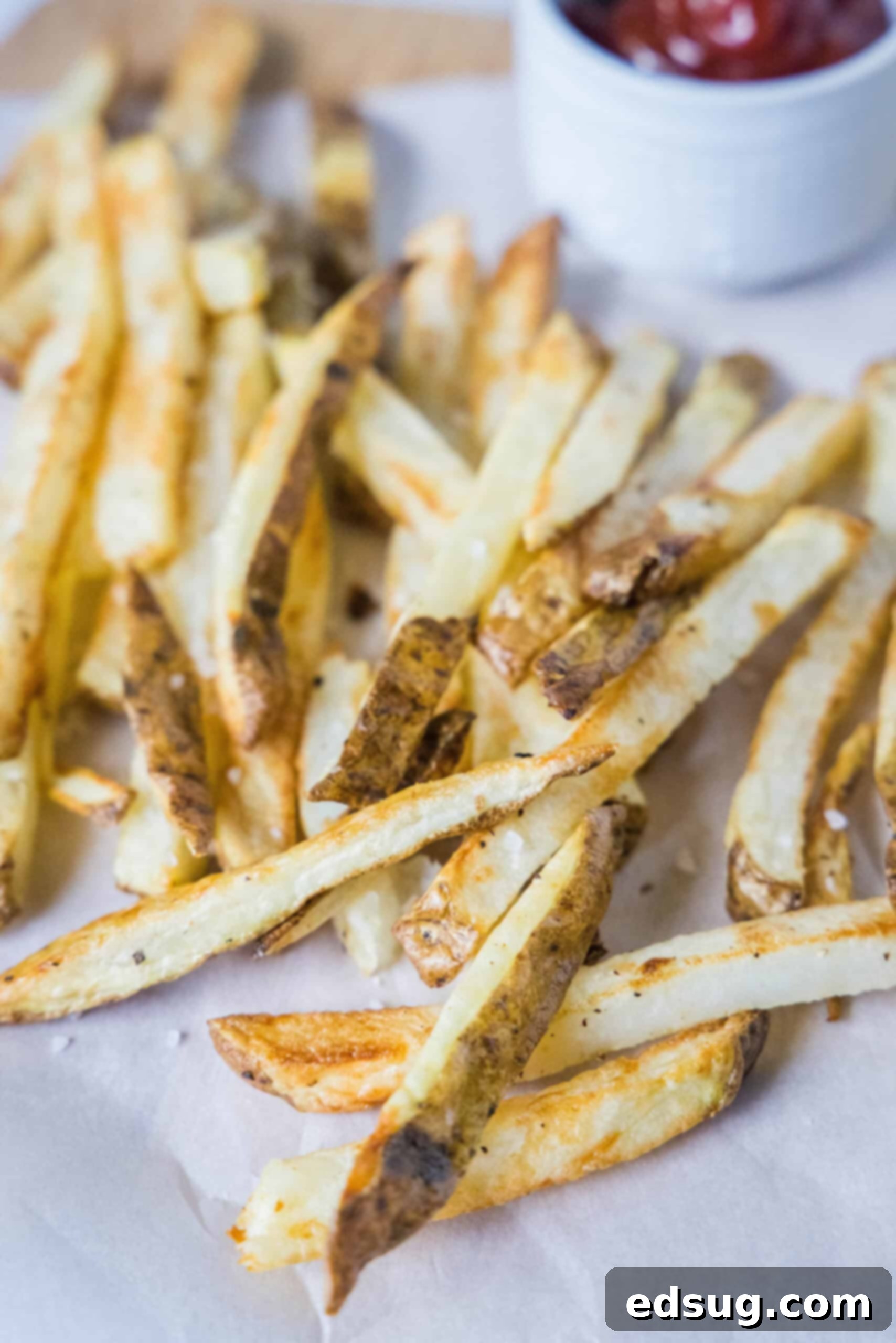 Homemade air fryer French fries are just as crispy as regular fries! Make them in a fraction of the time, with any seasonings you’d like. Homemade air fryer French fries on a sheet of parchment paper with a ramekin of ketchup in the background.