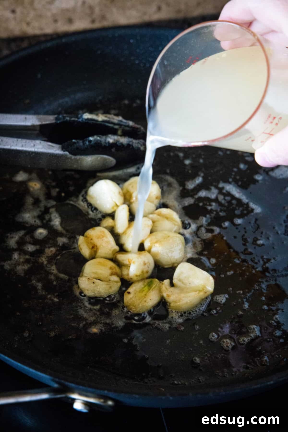 Chicken broth being poured into a skillet with sautéed garlic cloves.