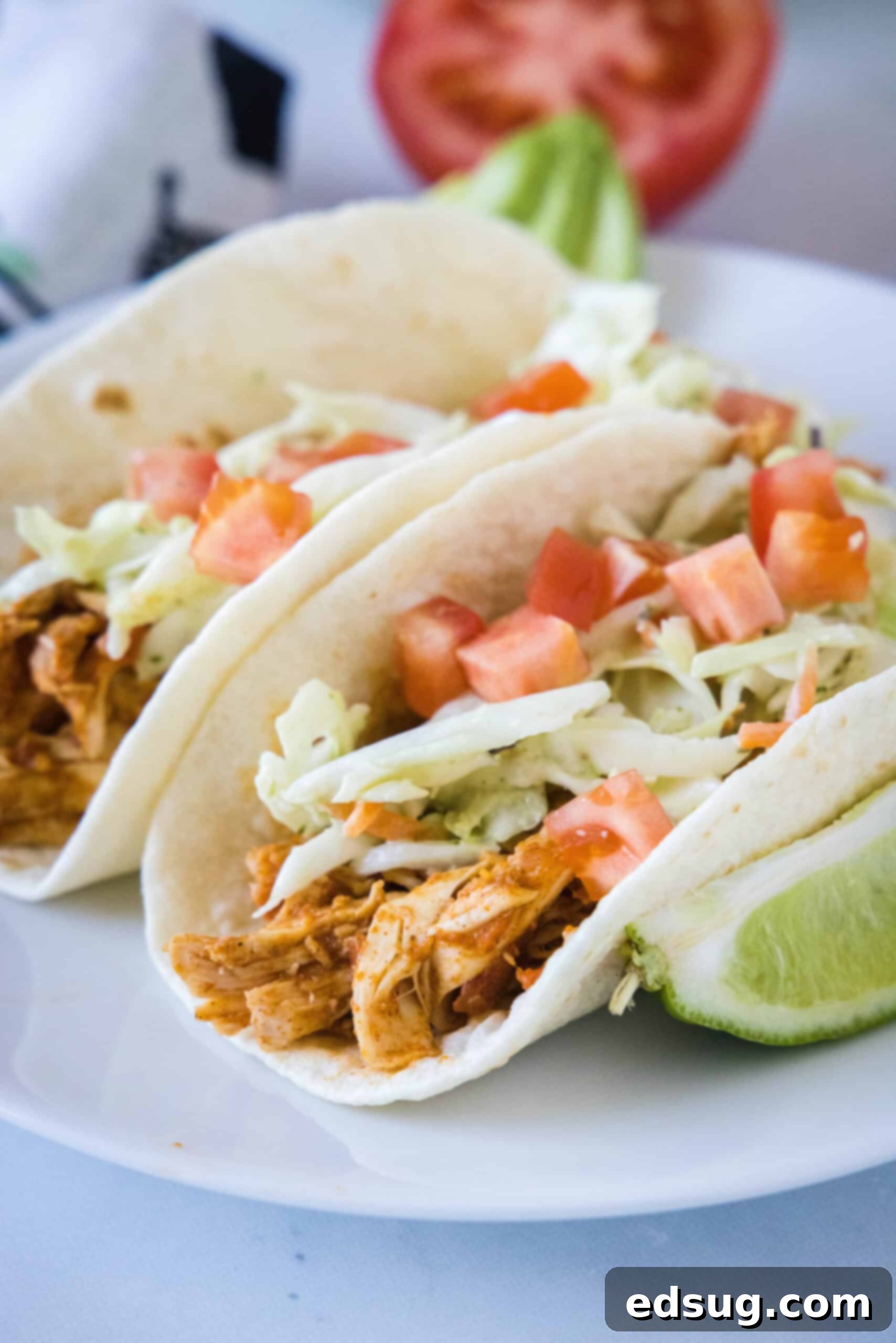 Two rotisserie chicken tacos topped with Mexican coleslaw and diced tomatoes on a white plate, next to a lime wedge.