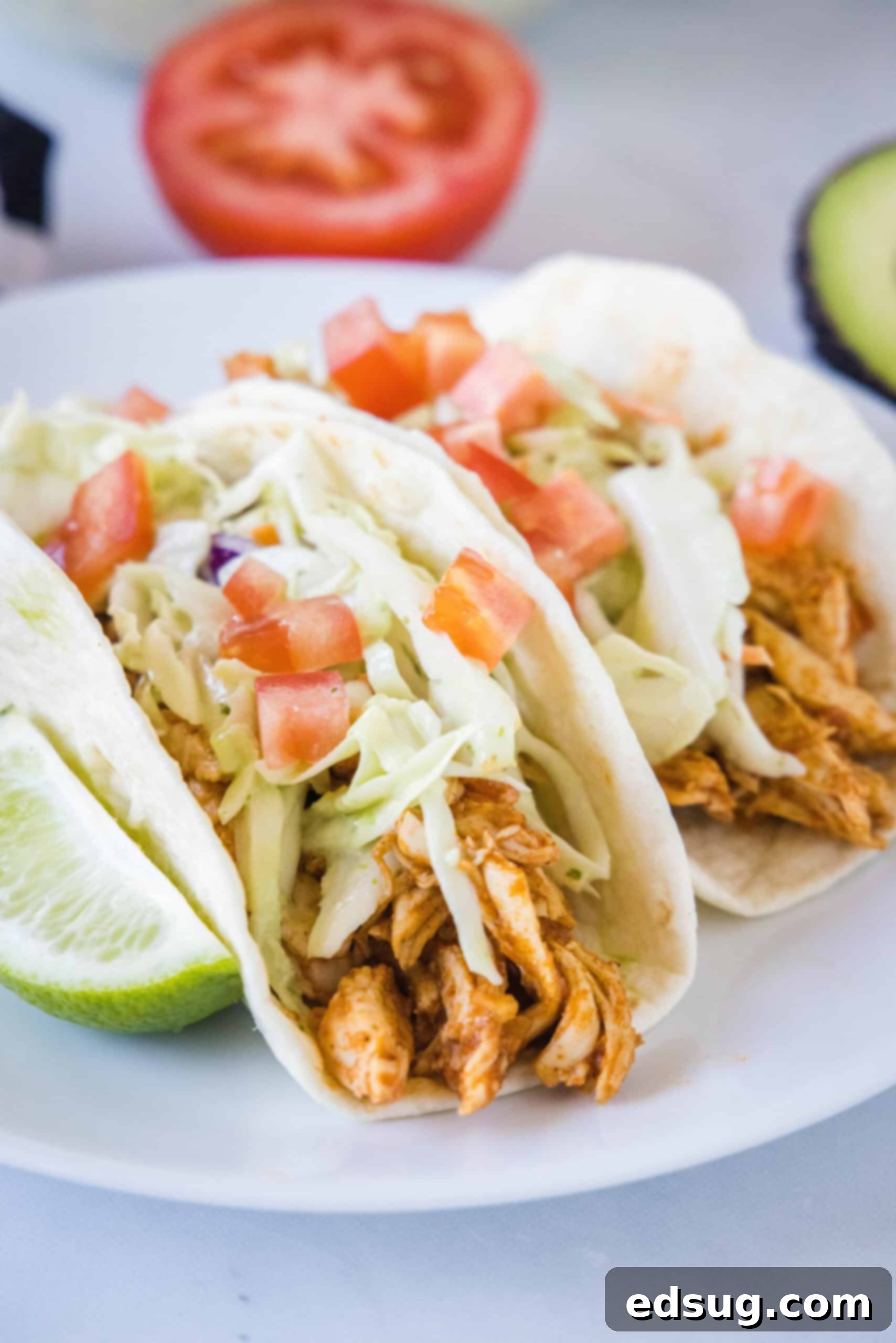Two rotisserie chicken tacos topped with Mexican coleslaw and diced tomatoes on a white plate, next to a lime wedge.