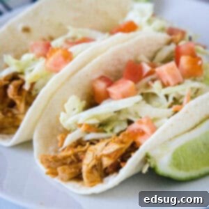 Two rotisserie chicken tacos topped with Mexican coleslaw and diced tomatoes on a white plate, next to a lime wedge.