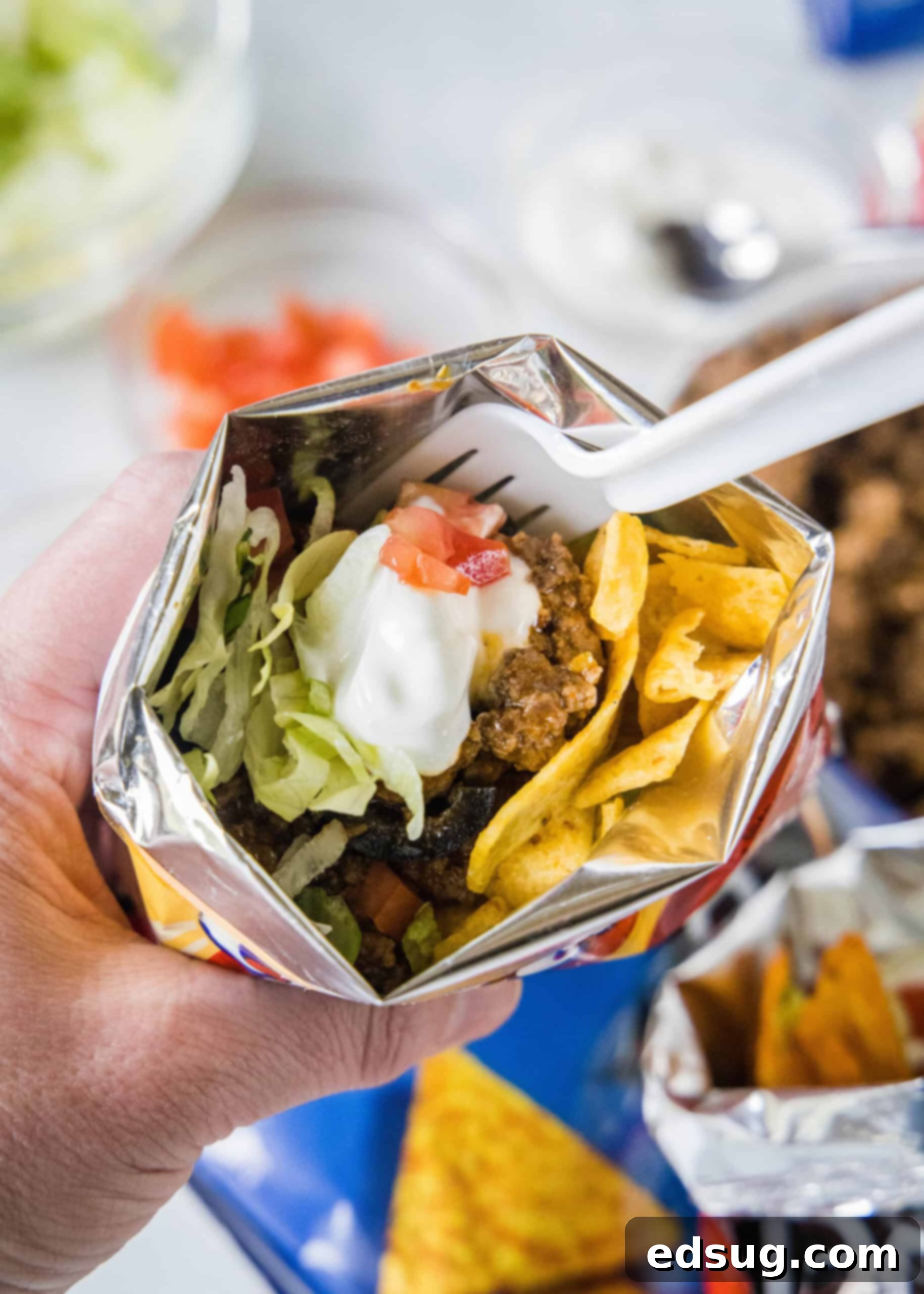 Enjoying a Handheld Walking Taco A hand holding a walking taco topped with crisp lettuce, fresh tomatoes, and creamy sour cream inside a chip bag, ready to be enjoyed with a fork.