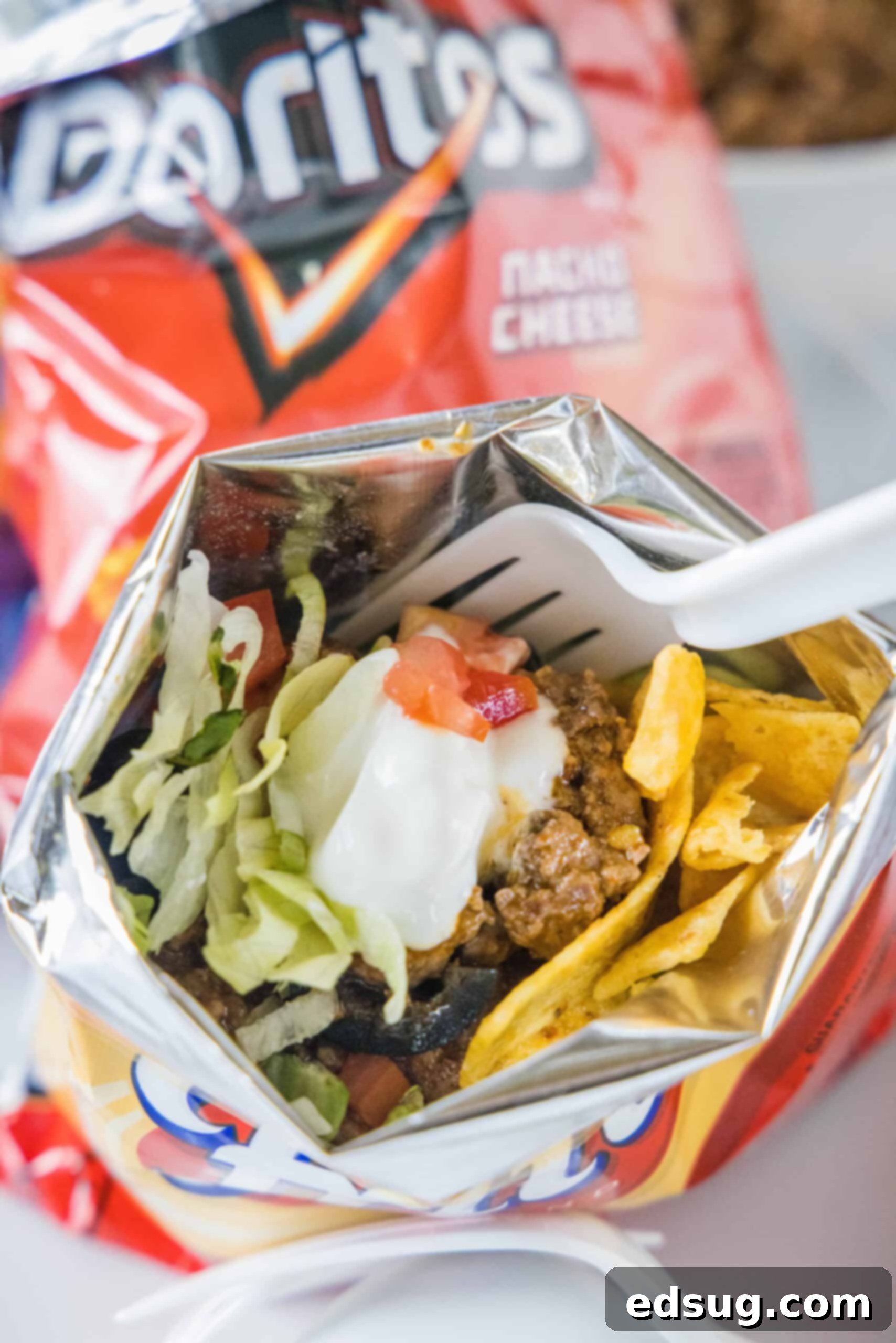 A Freshly Assembled Walking Taco A delicious walking taco topped with vibrant lettuce, diced tomatoes, and a dollop of sour cream, served inside a Fritos chip bag with a fork.