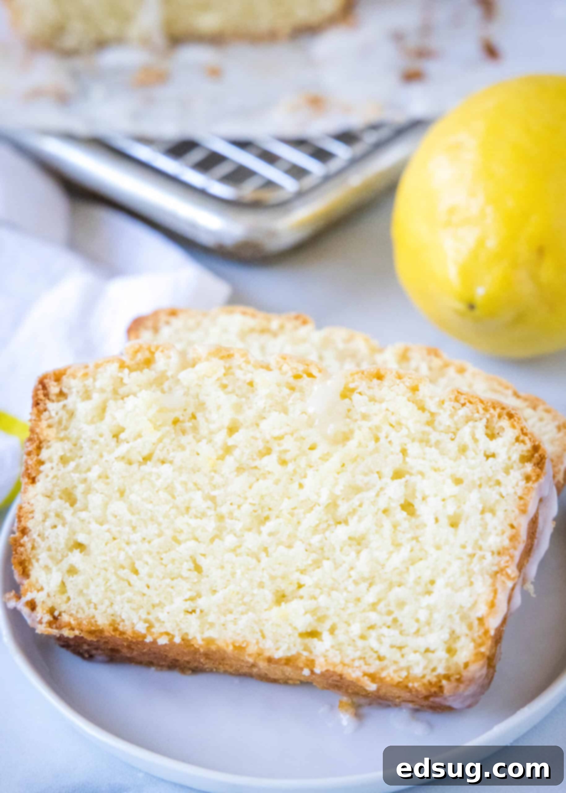 Two perfect slices of glazed lemon loaf on a white plate, with a fresh whole lemon in the background.