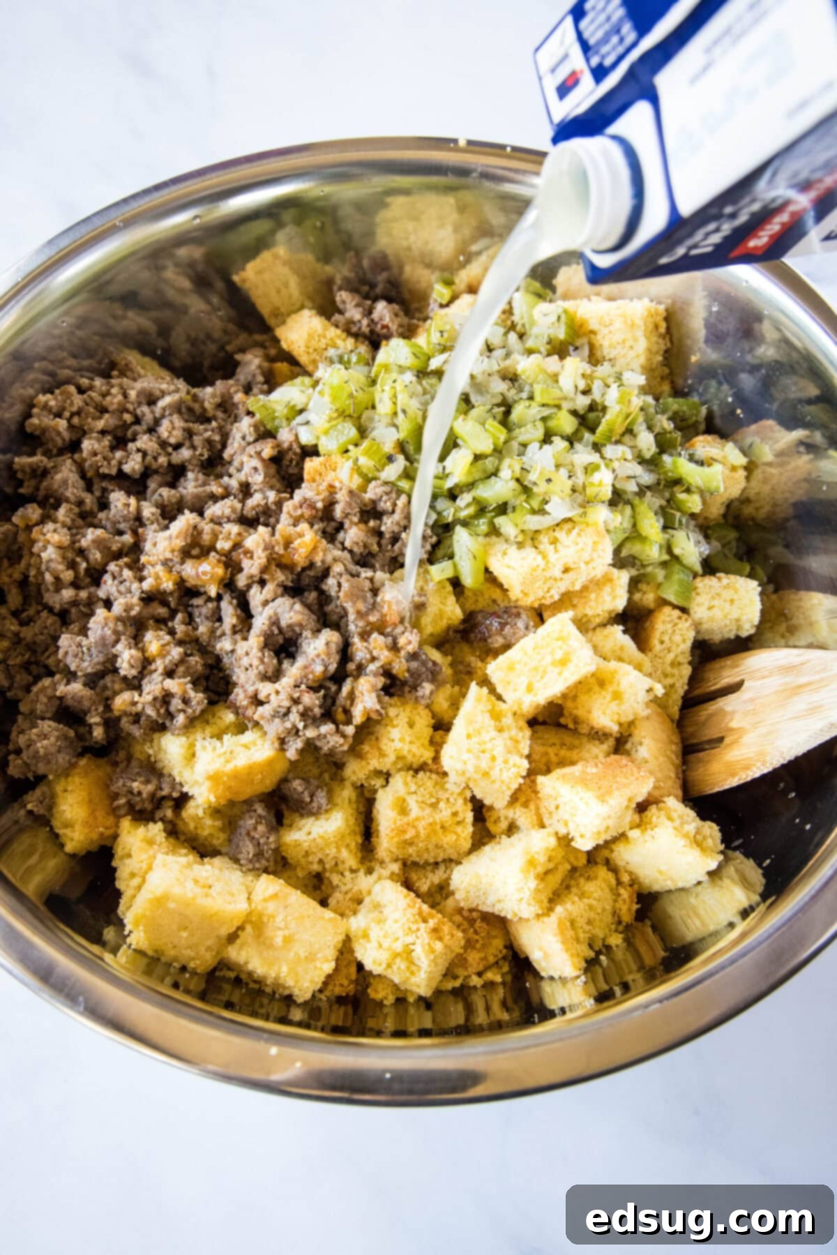 Chicken broth being poured into a mixing bowl with cornbread, sautéed onions and celery, and sausage meat.
