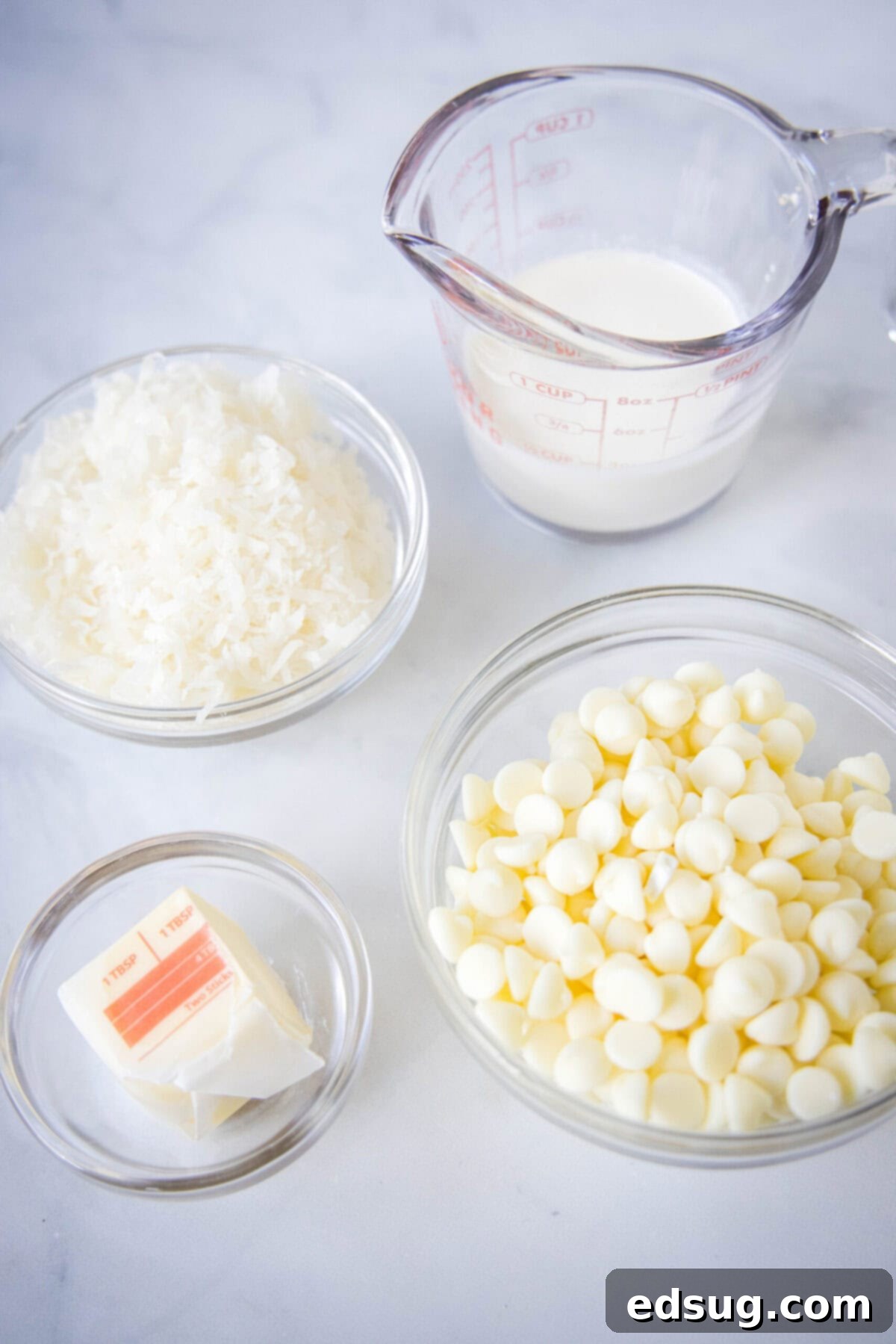 Overhead view of some of the ingredients needed for white chocolate coconut balls: a bowl of white chocolate chips, a pyrex of heavy cream, a bowl of shredded coconut, and some butter