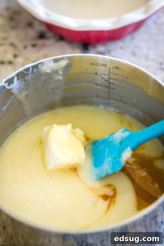 Softened butter and vanilla extract melting and mixing into the smooth white chocolate and cream ganache in a saucepan, being stirred with a rubber spatula