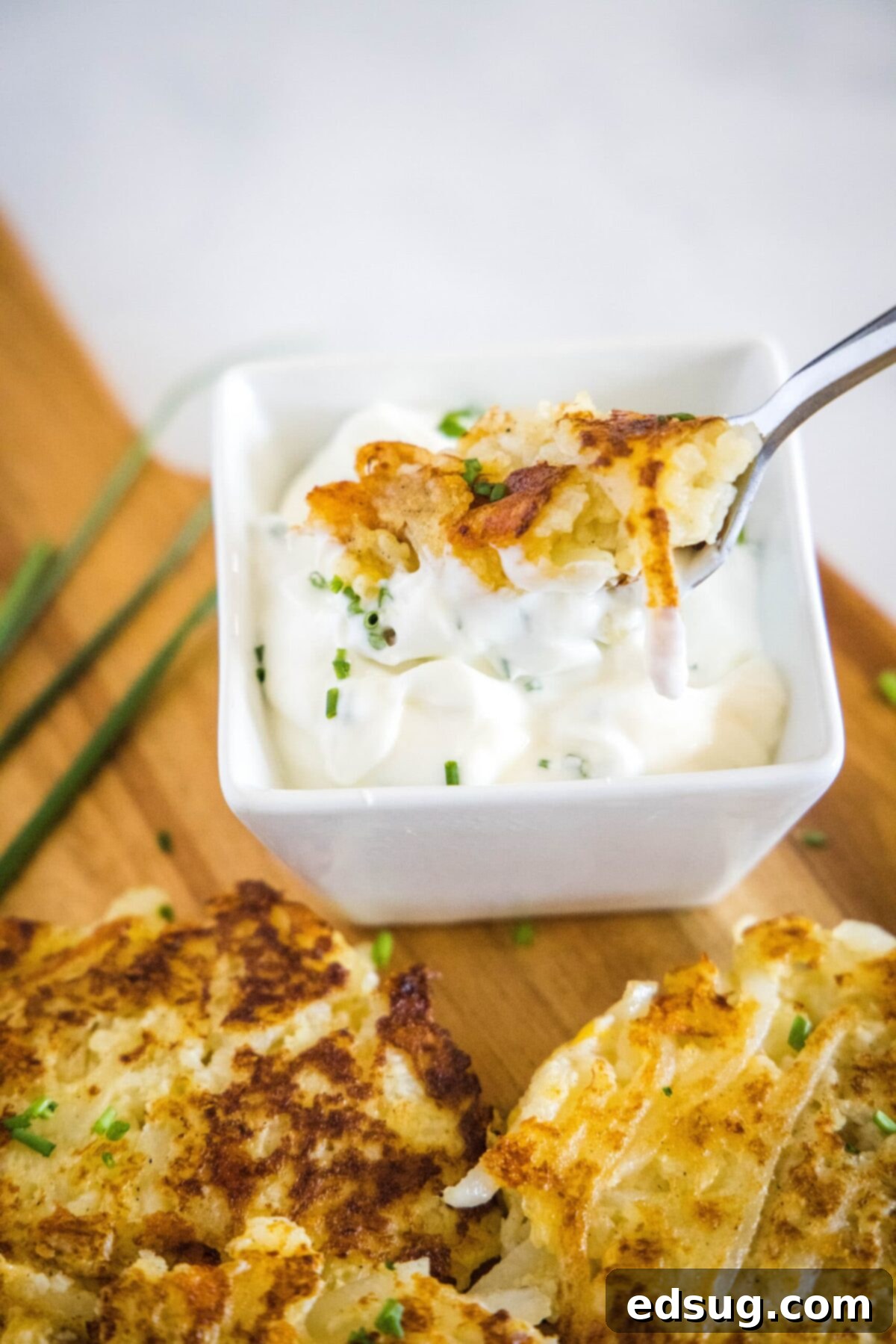 These easy cauliflower fritters are packed with hash browns and cheddar cheese, and served with a creamy and tart dipping sauce. A fork dipping a piece of cauliflower fritter into a bowl of sauce, on a cutting board with more fritters