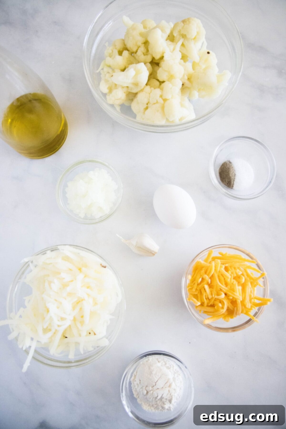These easy cauliflower fritters are packed with hash browns and cheddar cheese, and served with a creamy and tart dipping sauce. Overhead view of the ingredients needed to make cauliflower fritters: a bowl of cauliflower florets, a bowl of shredded hash browns, a bowl of grated cheddar cheese, a glass of olive oil, a bowl of salt and pepper, a bowl of chopped onion, a bowl of flour, a garlic clove, and an egg