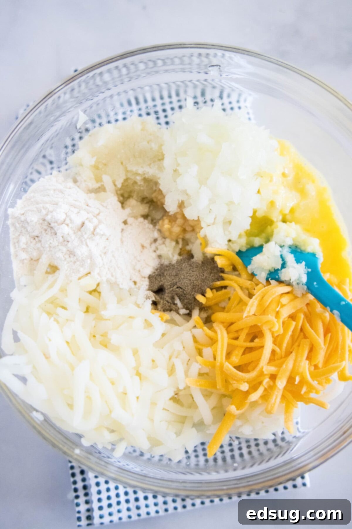 These easy cauliflower fritters are packed with hash browns and cheddar cheese, and served with a creamy and tart dipping sauce. A mixing bowl with mashed cauliflower, shredded hash browns, grated cheese, onion, garlic, salt, pepper, an egg, and a rubber spatula