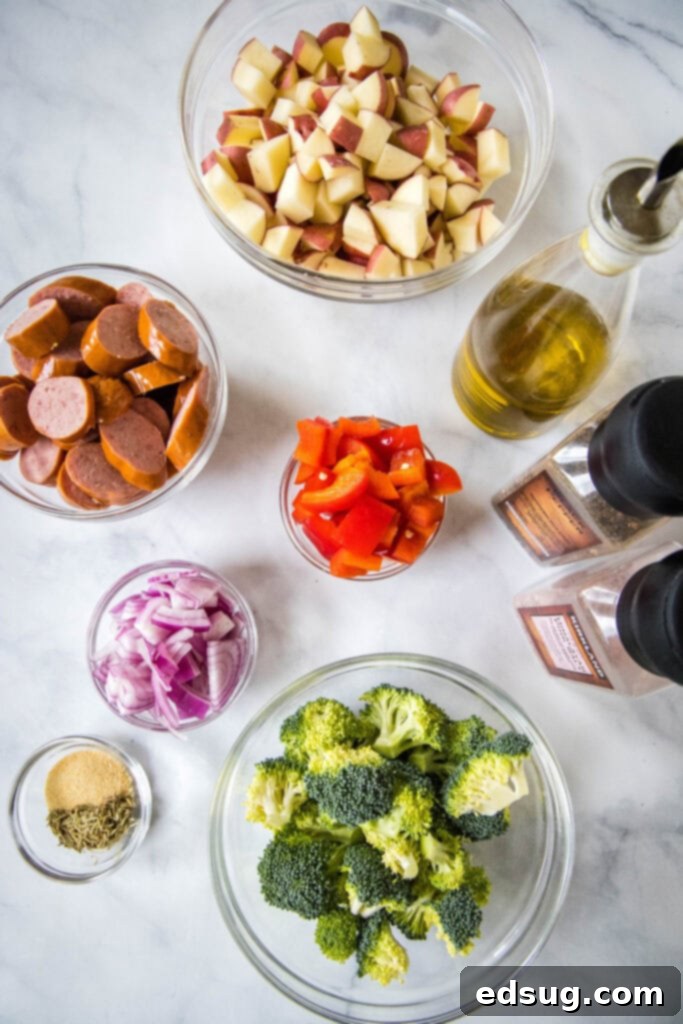 Flavorful One-Pan Sausage and Potatoes 3 A collection of fresh ingredients laid out, including red potatoes, a red onion, a red bell pepper, fresh broccoli florets, olive oil, and a package of sausage, ready for roasting.