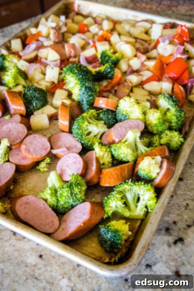 Flavorful One-Pan Sausage and Potatoes 5 A baking tray with partially roasted potatoes and vegetables, with fresh sliced sausage and broccoli florets being added to the empty space on the tray.